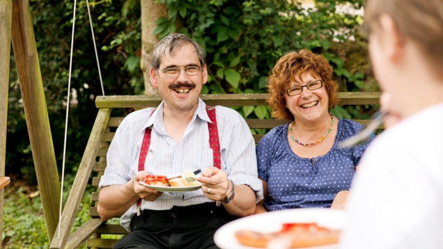 Ein Mann und eine Frau Essen auf einer Bank und lächeln einer gegenübersitzenden Person zu
