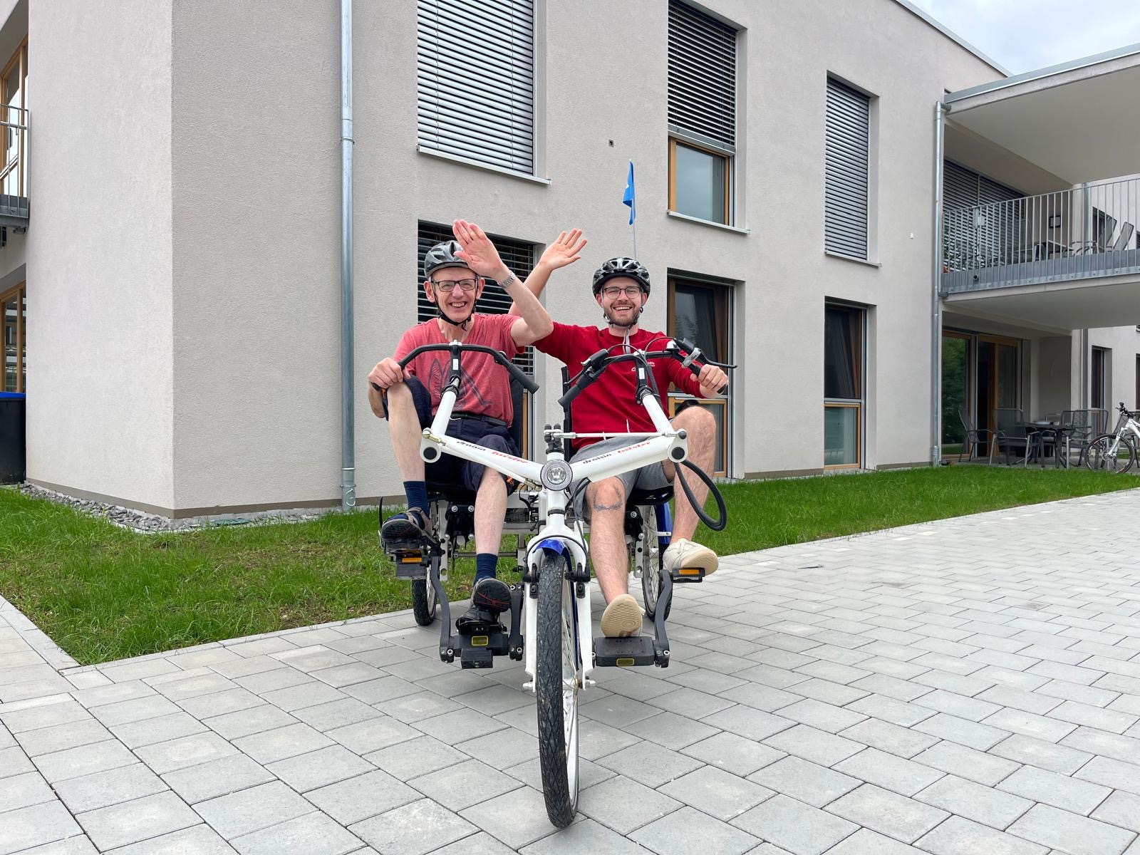 Zwei Menschen sitzen auf einem behindertengerechten Fahrrad und winken lächelnd